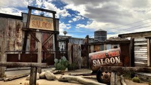 A vibrant dog park bathed in sunlight features abundant greenery with mature trees, offering ample shade for leisurely outdoor activities. In the distance is a well-equipped playground that presents an additional activity area. An inviting picnic table spreads out nearby, ideal for enjoying meals al fresco or taking relaxing breaks from play. A neatly-placed trash bin aids in maintaining the park's cleanliness for everyone's enjoyment. Dominating the scene is a generously wide dirt path gracefully curving through the expanse of grass, promising exploratory walks and lively fetch games.