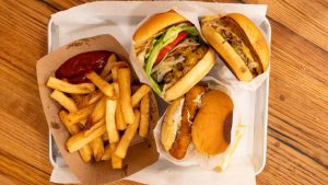 A vibrant dog park bathed in sunlight features abundant greenery with mature trees, offering ample shade for leisurely outdoor activities. In the distance is a well-equipped playground that presents an additional activity area. An inviting picnic table spreads out nearby, ideal for enjoying meals al fresco or taking relaxing breaks from play. A neatly-placed trash bin aids in maintaining the park's cleanliness for everyone's enjoyment. Dominating the scene is a generously wide dirt path gracefully curving through the expanse of grass, promising exploratory walks and lively fetch games.