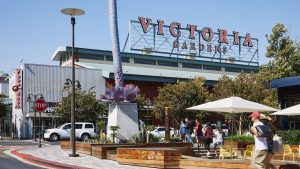 A vibrant dog park bathed in sunlight features abundant greenery with mature trees, offering ample shade for leisurely outdoor activities. In the distance is a well-equipped playground that presents an additional activity area. An inviting picnic table spreads out nearby, ideal for enjoying meals al fresco or taking relaxing breaks from play. A neatly-placed trash bin aids in maintaining the park's cleanliness for everyone's enjoyment. Dominating the scene is a generously wide dirt path gracefully curving through the expanse of grass, promising exploratory walks and lively fetch games.