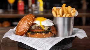 A vibrant dog park bathed in sunlight features abundant greenery with mature trees, offering ample shade for leisurely outdoor activities. In the distance is a well-equipped playground that presents an additional activity area. An inviting picnic table spreads out nearby, ideal for enjoying meals al fresco or taking relaxing breaks from play. A neatly-placed trash bin aids in maintaining the park's cleanliness for everyone's enjoyment. Dominating the scene is a generously wide dirt path gracefully curving through the expanse of grass, promising exploratory walks and lively fetch games.