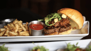 A vibrant dog park bathed in sunlight features abundant greenery with mature trees, offering ample shade for leisurely outdoor activities. In the distance is a well-equipped playground that presents an additional activity area. An inviting picnic table spreads out nearby, ideal for enjoying meals al fresco or taking relaxing breaks from play. A neatly-placed trash bin aids in maintaining the park's cleanliness for everyone's enjoyment. Dominating the scene is a generously wide dirt path gracefully curving through the expanse of grass, promising exploratory walks and lively fetch games.