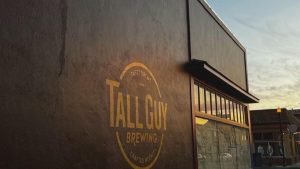 A vibrant dog park bathed in sunlight features abundant greenery with mature trees, offering ample shade for leisurely outdoor activities. In the distance is a well-equipped playground that presents an additional activity area. An inviting picnic table spreads out nearby, ideal for enjoying meals al fresco or taking relaxing breaks from play. A neatly-placed trash bin aids in maintaining the park's cleanliness for everyone's enjoyment. Dominating the scene is a generously wide dirt path gracefully curving through the expanse of grass, promising exploratory walks and lively fetch games.