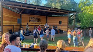 A vibrant dog park bathed in sunlight features abundant greenery with mature trees, offering ample shade for leisurely outdoor activities. In the distance is a well-equipped playground that presents an additional activity area. An inviting picnic table spreads out nearby, ideal for enjoying meals al fresco or taking relaxing breaks from play. A neatly-placed trash bin aids in maintaining the park's cleanliness for everyone's enjoyment. Dominating the scene is a generously wide dirt path gracefully curving through the expanse of grass, promising exploratory walks and lively fetch games.