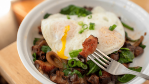 A vibrant dog park bathed in sunlight features abundant greenery with mature trees, offering ample shade for leisurely outdoor activities. In the distance is a well-equipped playground that presents an additional activity area. An inviting picnic table spreads out nearby, ideal for enjoying meals al fresco or taking relaxing breaks from play. A neatly-placed trash bin aids in maintaining the park's cleanliness for everyone's enjoyment. Dominating the scene is a generously wide dirt path gracefully curving through the expanse of grass, promising exploratory walks and lively fetch games.