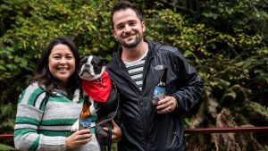 A vibrant dog park bathed in sunlight features abundant greenery with mature trees, offering ample shade for leisurely outdoor activities. In the distance is a well-equipped playground that presents an additional activity area. An inviting picnic table spreads out nearby, ideal for enjoying meals al fresco or taking relaxing breaks from play. A neatly-placed trash bin aids in maintaining the park's cleanliness for everyone's enjoyment. Dominating the scene is a generously wide dirt path gracefully curving through the expanse of grass, promising exploratory walks and lively fetch games.