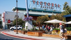 A vibrant dog park bathed in sunlight features abundant greenery with mature trees, offering ample shade for leisurely outdoor activities. In the distance is a well-equipped playground that presents an additional activity area. An inviting picnic table spreads out nearby, ideal for enjoying meals al fresco or taking relaxing breaks from play. A neatly-placed trash bin aids in maintaining the park's cleanliness for everyone's enjoyment. Dominating the scene is a generously wide dirt path gracefully curving through the expanse of grass, promising exploratory walks and lively fetch games.