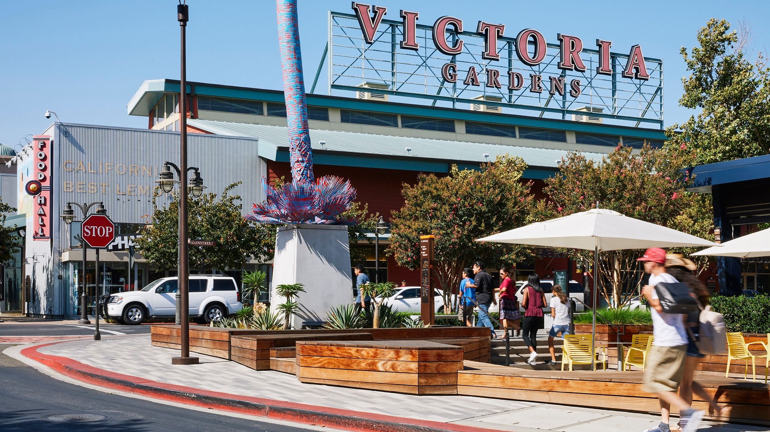 A vibrant dog park bathed in sunlight features abundant greenery with mature trees, offering ample shade for leisurely outdoor activities. In the distance is a well-equipped playground that presents an additional activity area. An inviting picnic table spreads out nearby, ideal for enjoying meals al fresco or taking relaxing breaks from play. A neatly-placed trash bin aids in maintaining the park's cleanliness for everyone's enjoyment. Dominating the scene is a generously wide dirt path gracefully curving through the expanse of grass, promising exploratory walks and lively fetch games.